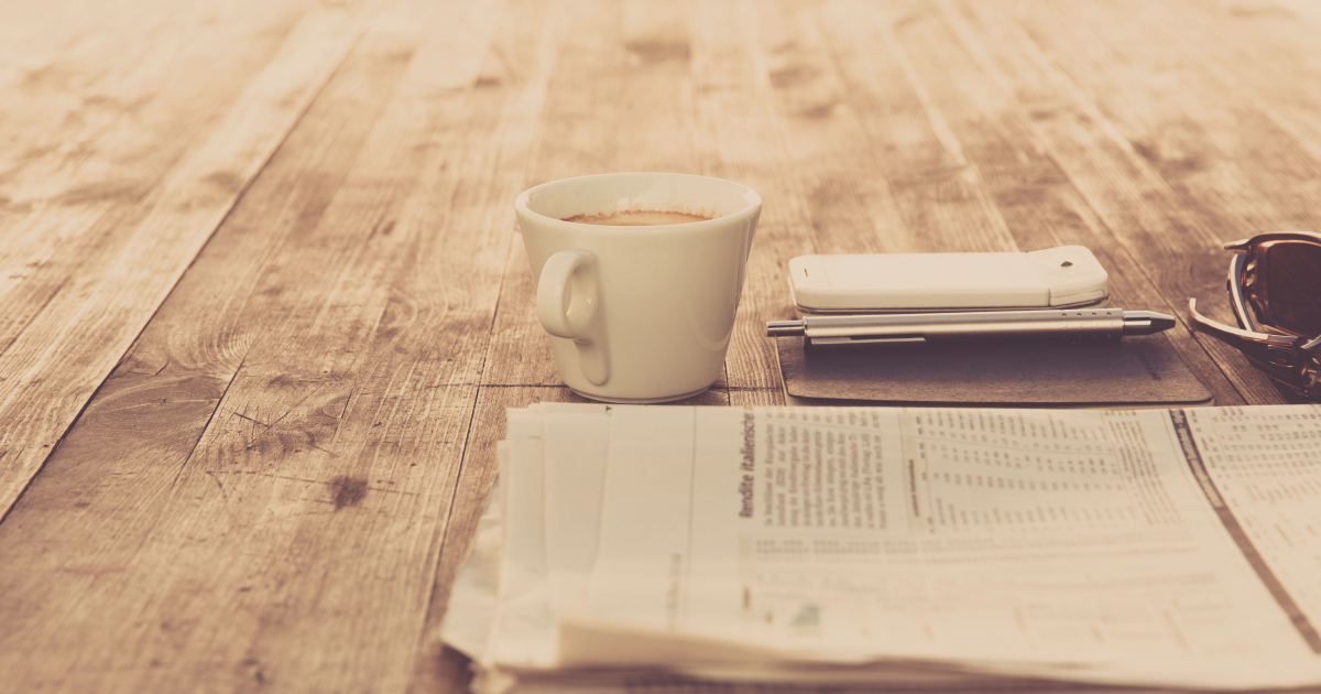 A photo of a wooden table with a newspaper and a cup of coffee. 